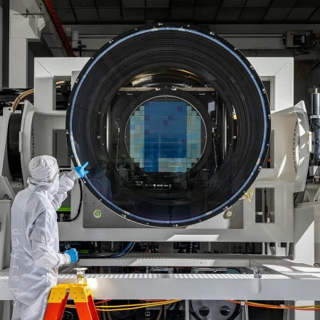 Scientist in clean suit points at a large blue wafer inside of a black lens of a white machine in a bright research lab.