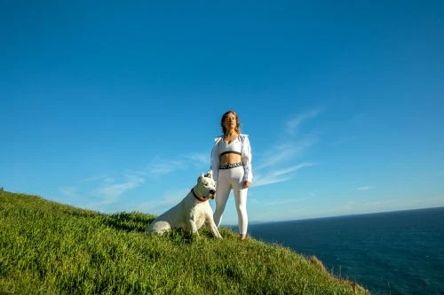 Woman in athletic wear poses with a large white dog on a grassy hill against a blue sky and ocean backdrop in a wide shot.