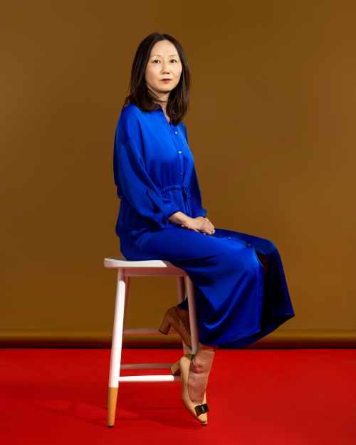 Elegant Asian woman in a royal blue dress poses sitting on a stool against a warm brown backdrop with a red floor.