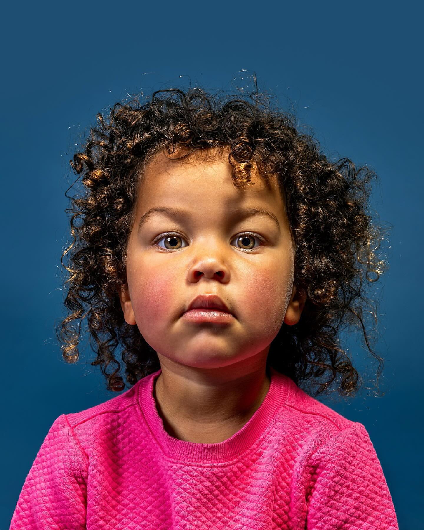 Portrait of a young child with curly hair, wearing a pink sweatshirt, against a dark blue background.