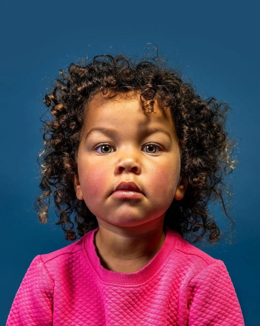 Portrait of a young child with curly hair, wearing a pink sweatshirt, against a dark blue background.