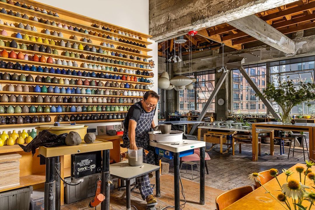 Adult male artist shapes clay on a pottery wheel in a studio filled with colorful ceramic pieces and natural light from large windows.