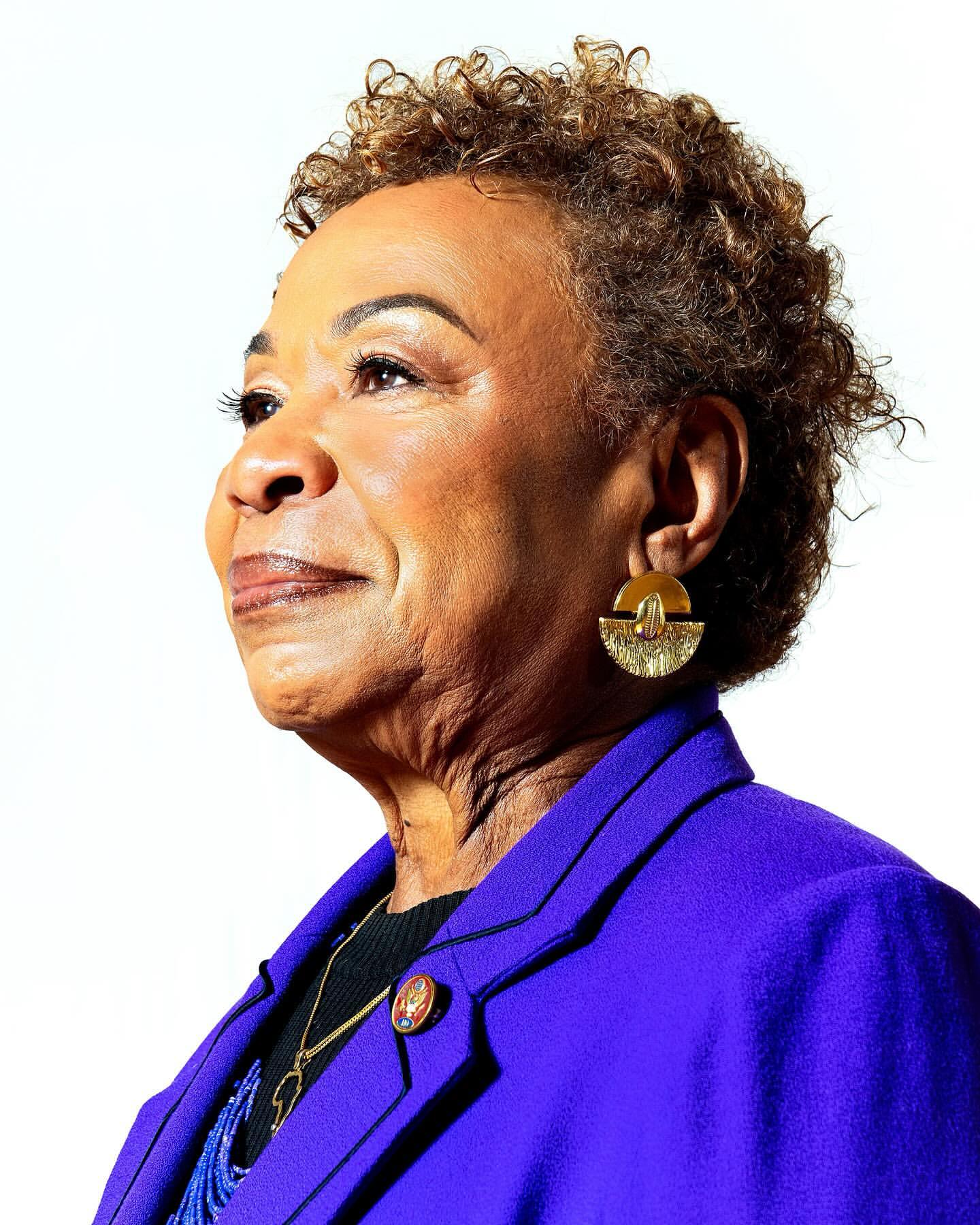 Close-up portrait of an adult woman with short curly hair, wearing a purple blazer, gold earrings, and looking towards the upper left.