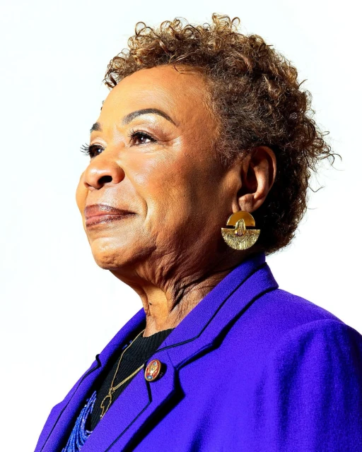 Close-up portrait of an adult woman with short curly hair, wearing a purple blazer, gold earrings, and looking towards the upper left.