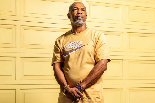 Portrait of a grey-haired man in a gold-colored Nike t-shirt standing against a yellow paneled wall, looking upwards thoughtfully.