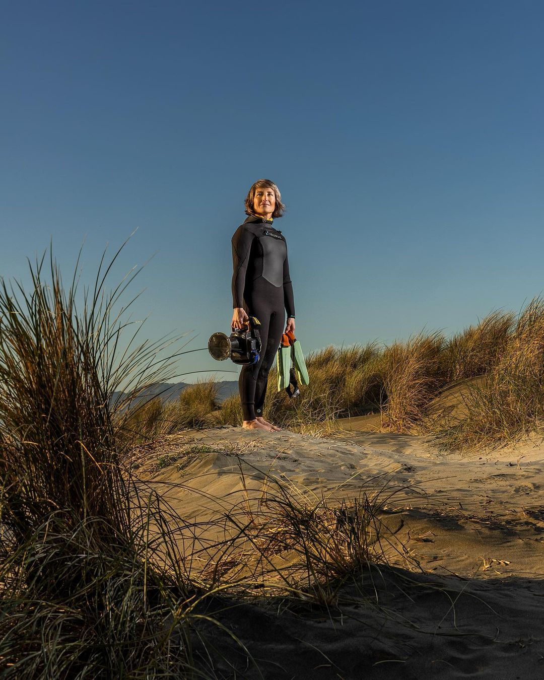 Full-length portrait of a woman in a wetsuit standing on a dune with dunes and clear blue sky in the background, holding a camera.