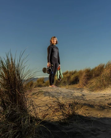 Full-length portrait of a woman in a wetsuit standing on a dune with dunes and clear blue sky in the background, holding a camera.