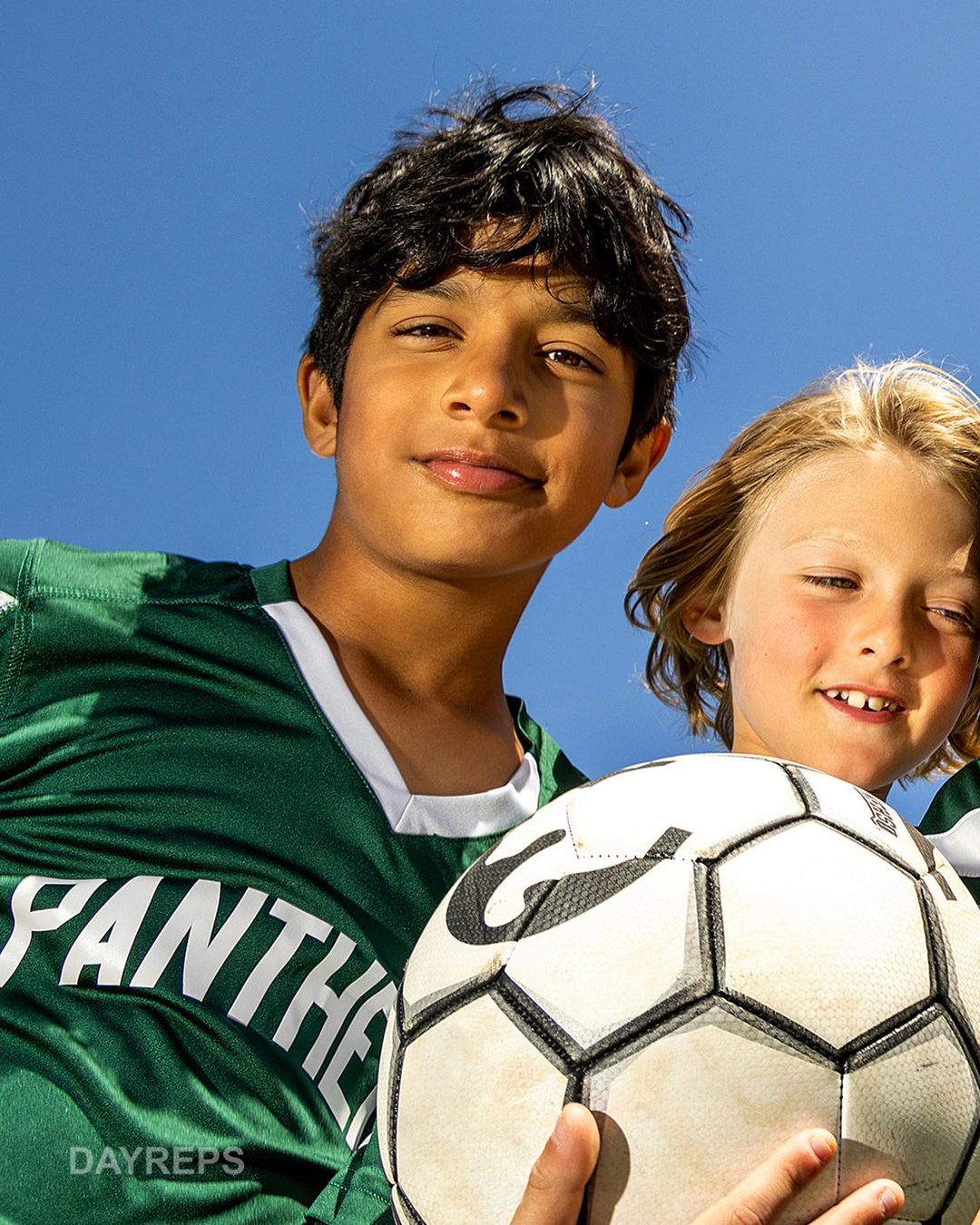 Two smiling kids in soccer jerseys hold a soccer ball in front of a clear blue sky. Close-up shot showcases their youthful energy.