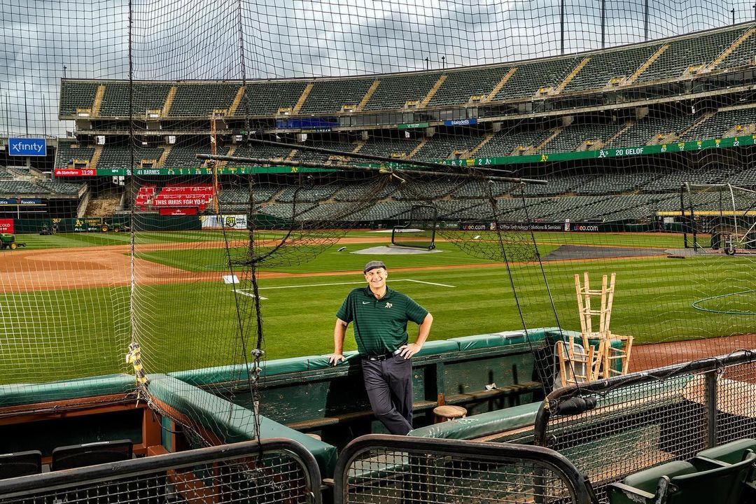 Man leans against dugout in baseball stadium, wearing a green polo shirt and cap, with empty green seats and dark sky visible.