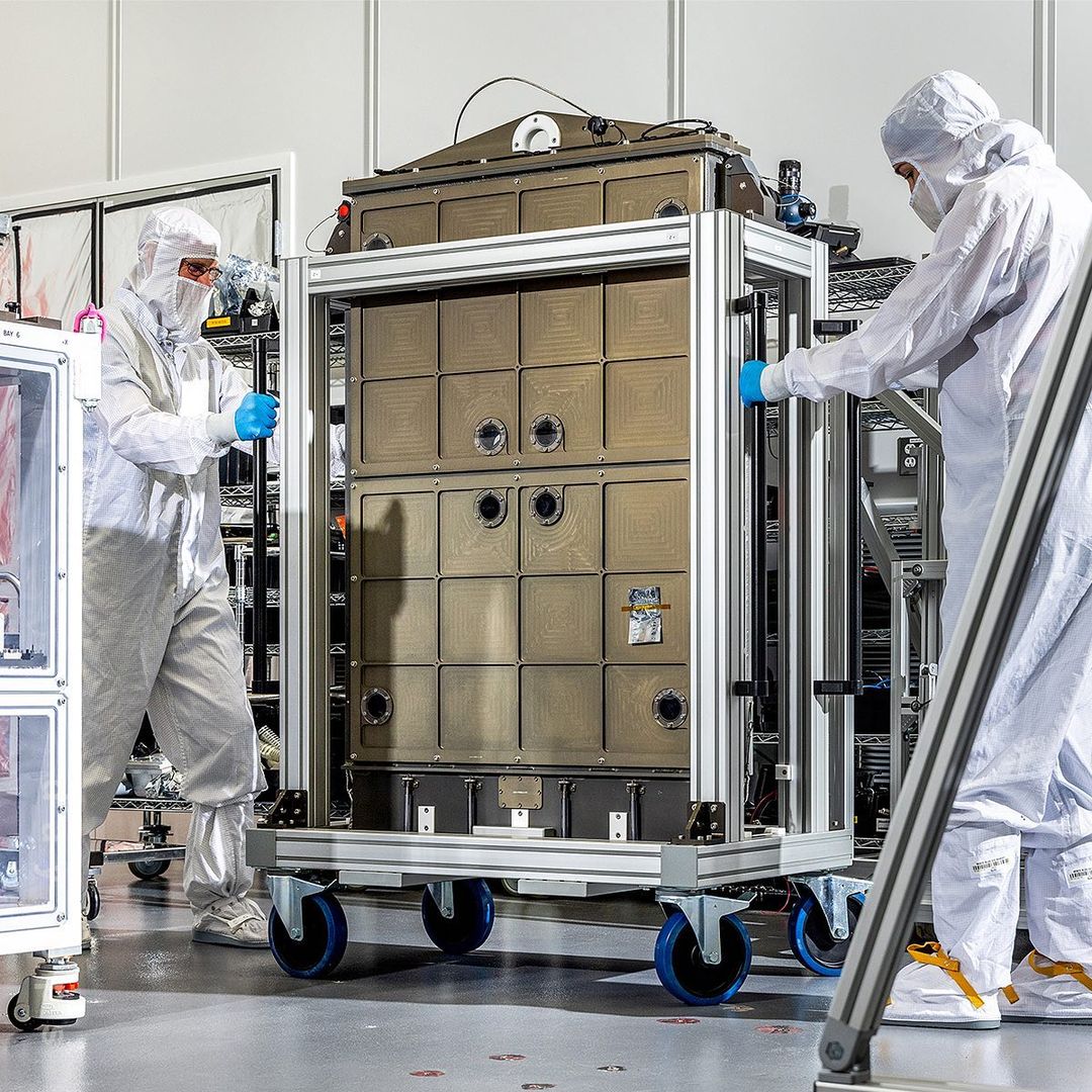 Two people in clean suits maneuvering a large, box-shaped metal container on a wheeled platform in a cleanroom environment.