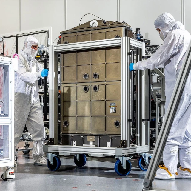 Two people in clean suits maneuvering a large, box-shaped metal container on a wheeled platform in a cleanroom environment.