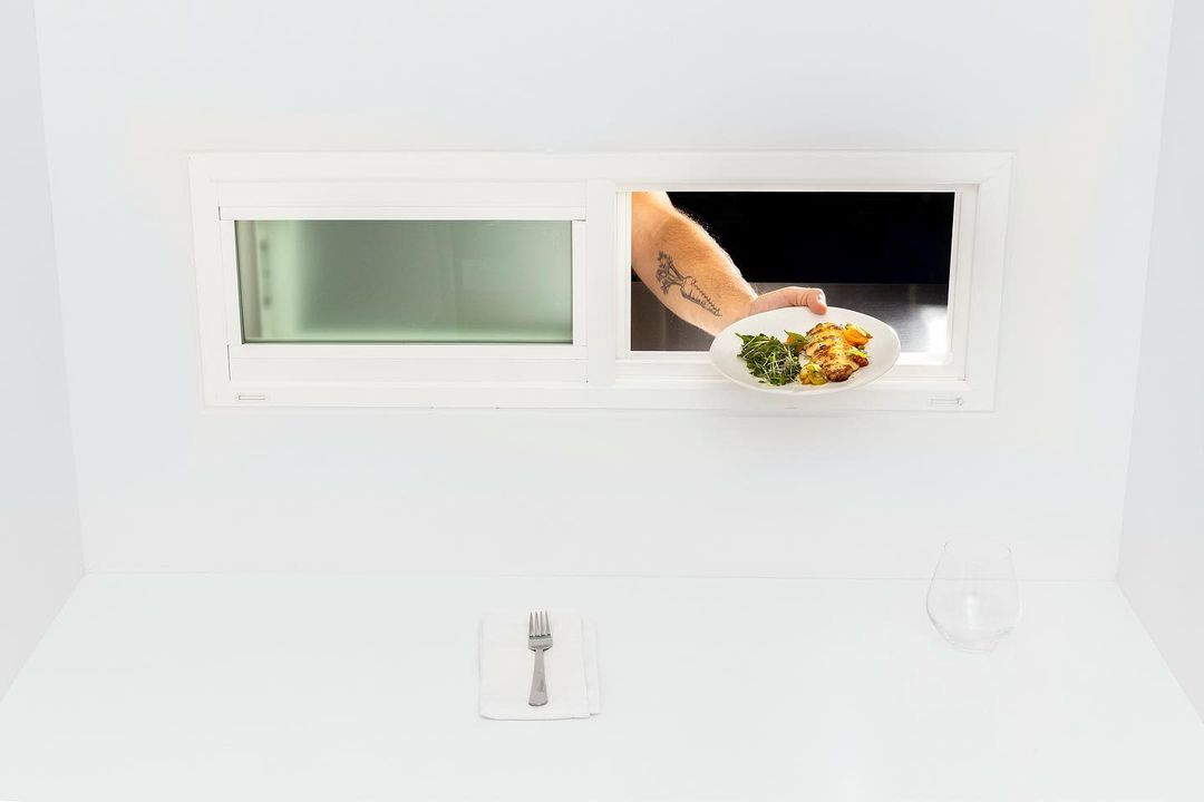 Arm holds plate of prepared food through white window frame above table setting with napkin, silverware and glass in minimal space.