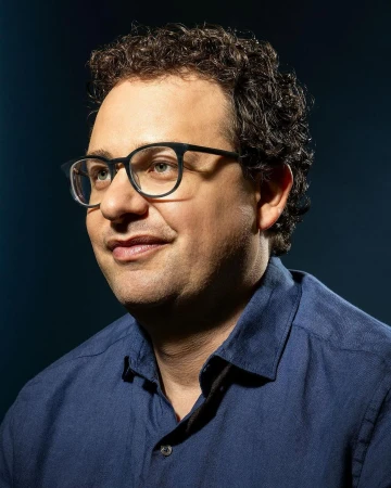 Close-up portrait of a man in his 40s wearing glasses and a blue shirt against a dark textured background