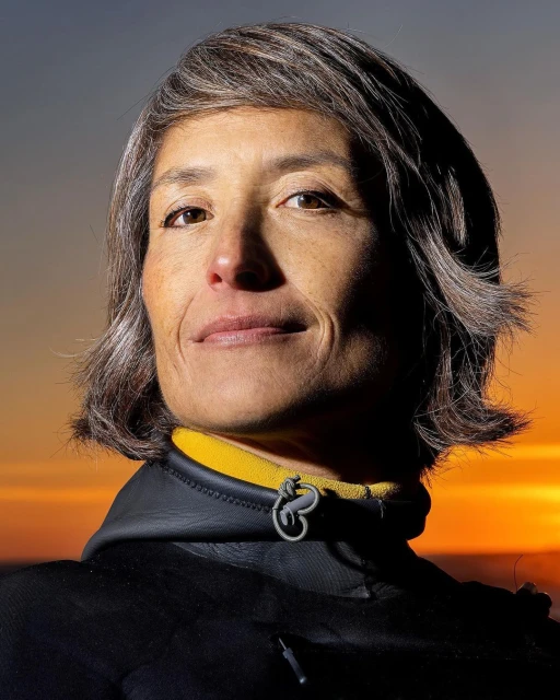 Portrait of an adult woman with short gray hair, wearing a black wetsuit with a yellow collar against a sunset background.