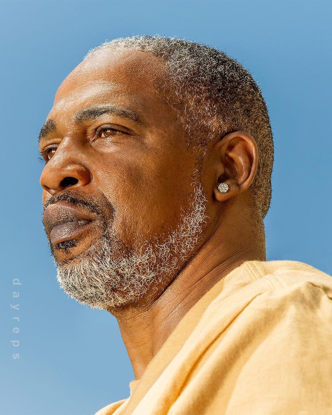Portrait of an African American man with grey hair and beard against a clear blue sky, wearing a yellow shirt and diamond earring.