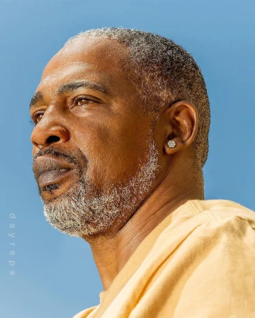 Portrait of an African American man with grey hair and beard against a clear blue sky, wearing a yellow shirt and diamond earring.