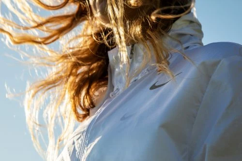 A person wearing a white Nike windbreaker with golden hair blowing in the wind against a clear blue sky, captured in an outdoor setting.