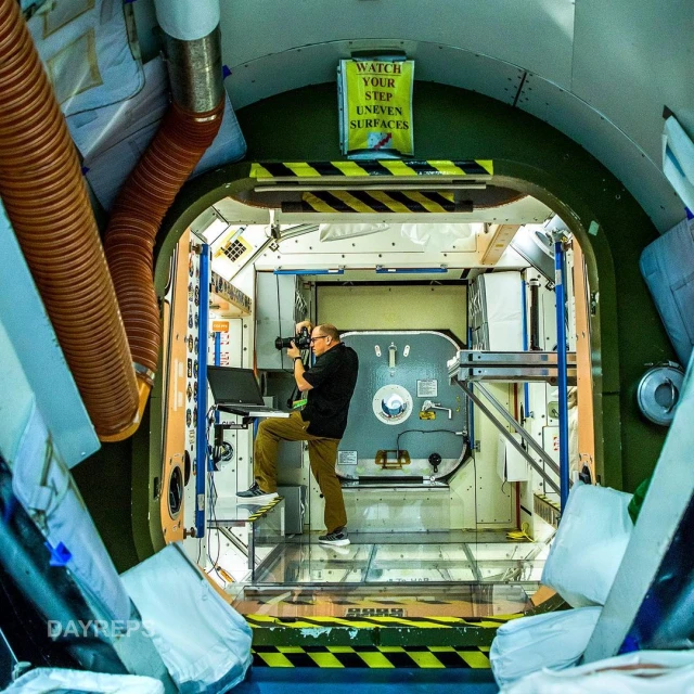 Inside a space station module, an adult male photographs the interior while standing on a small platform near a laptop, illuminated by interior lighting.