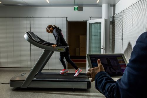 Woman in workout clothing uses a treadmill indoors while being monitored. Modern building with white walls, indoor exercise setting.
