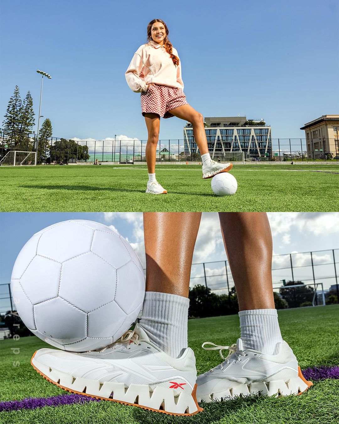 Female athlete in sportswear stands with foot on white soccer ball on green sports field under blue sky with buildings in the distance.