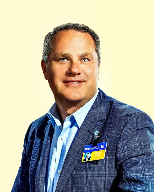 Headshot of an adult male in a suit and collared shirt with a name tag, posing in front of a soft yellow backdrop in a studio setting.