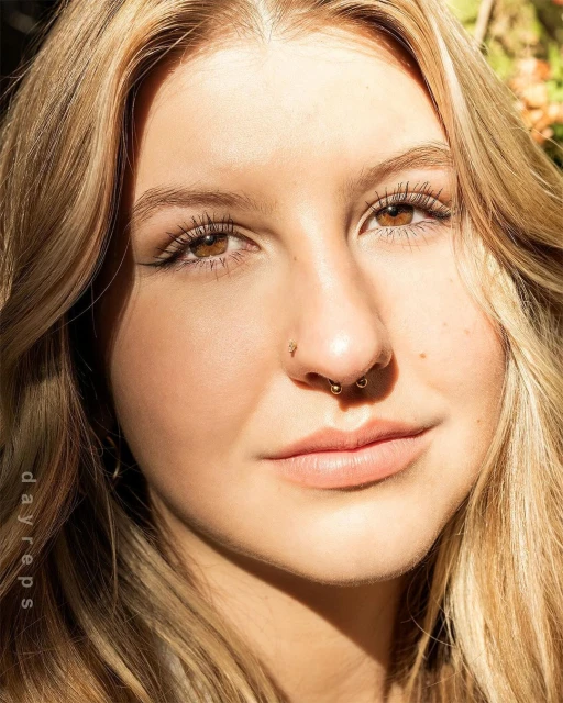 Close-up portrait of a young adult woman with blonde hair, brown eyes, and nose piercings smiling in natural outdoor lighting.