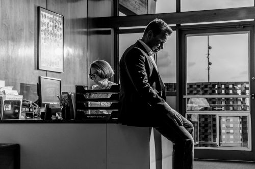 Monochrome shot of a businessman sitting pensively on a windowsill, with a woman working at her desk in the background.