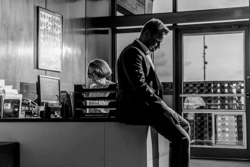 Monochrome shot of a businessman sitting pensively on a windowsill, with a woman working at her desk in the background.
