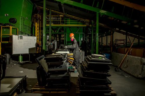 Worker in safety gear operates computer amid stacked electronics in recycling plant, under overhead conveyor system and green industrial machinery.