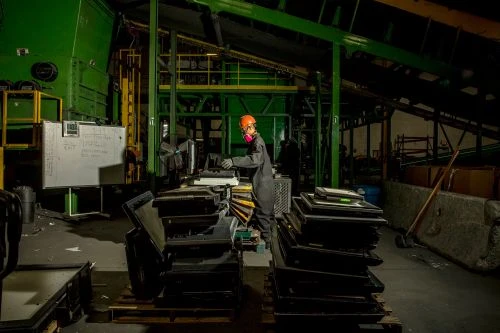 Worker in safety gear operates computer amid stacked electronics in recycling plant, under overhead conveyor system and green industrial machinery.