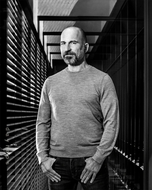 Monochrome portrait of a man with a beard, wearing a grey sweater and jeans, hands in pockets, posing against a modern architectural backdrop.