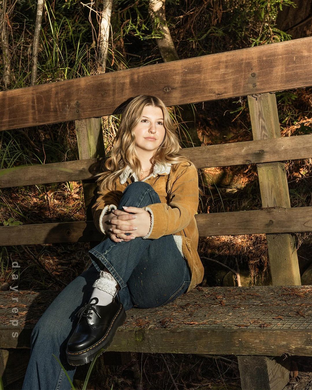 A young woman with blonde hair sits on a wooden bench in a wooded area, wearing a corduroy jacket and jeans, bathed in natural light.