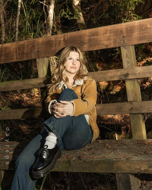 A young woman with blonde hair sits on a wooden bench in a wooded area, wearing a corduroy jacket and jeans, bathed in natural light.