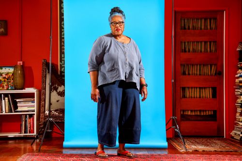 Full shot of a mature woman with blue hair wearing casual blue separates, posing in front of a blue backdrop with red walls and a door.