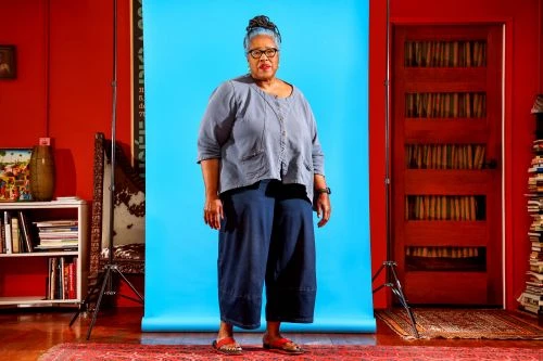 Full shot of a mature woman with blue hair wearing casual blue separates, posing in front of a blue backdrop with red walls and a door.