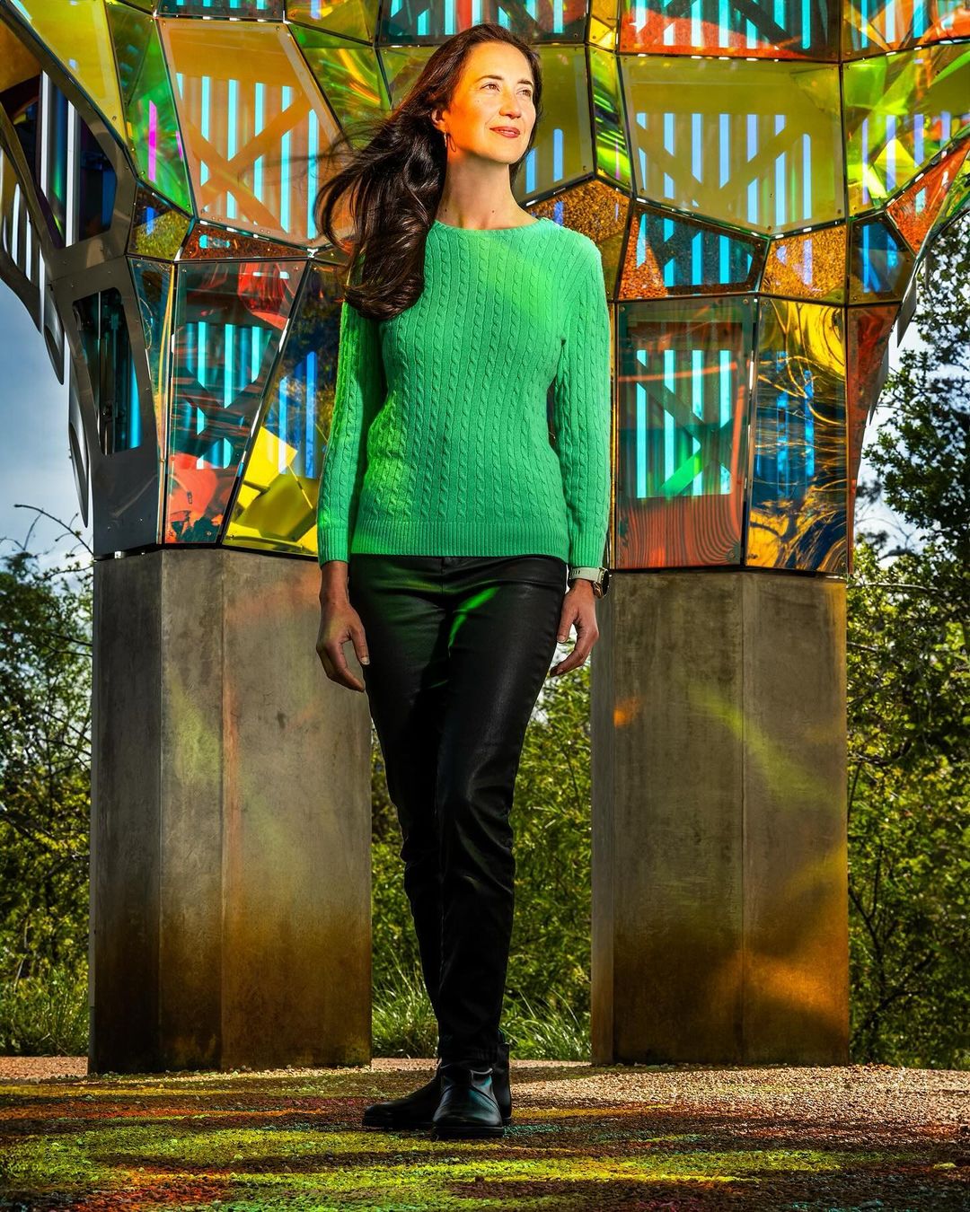 Attractive brunette woman in a green sweater and black pants poses next to colorful modern art in a park setting with bright sunlight.