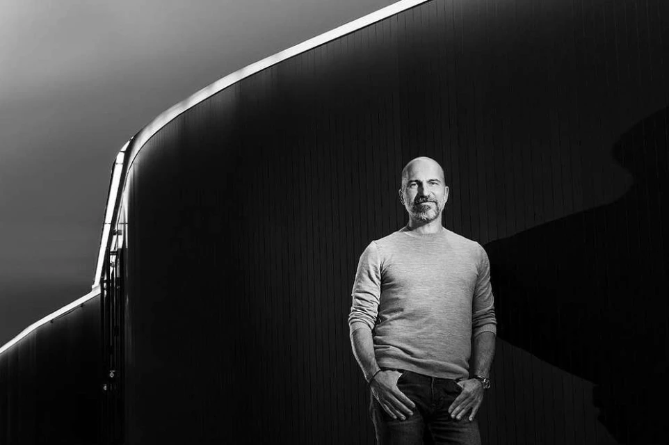 Monochrome portrait of a bald adult male with a beard, wearing a sweater and jeans, posing outdoors against an architectural wall.