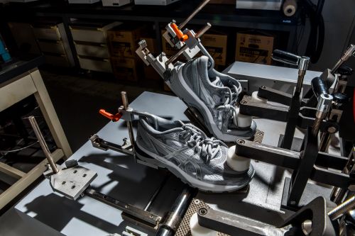 Two grey athletic shoes are secured in a metal testing apparatus in a lab setting, prepared for stress or durability testing.