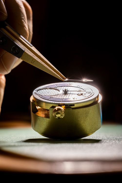 Close-up of a watchmaker's hands using tweezers to adjust a watch mechanism with the watch on a green surface in a dark room.