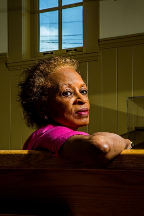 Mature Black woman with short hair wearing pink top, sitting in church pew with window in background looking toward the viewer.