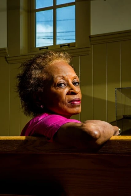 Mature Black woman with short hair wearing pink top, sitting in church pew with window in background looking toward the viewer.