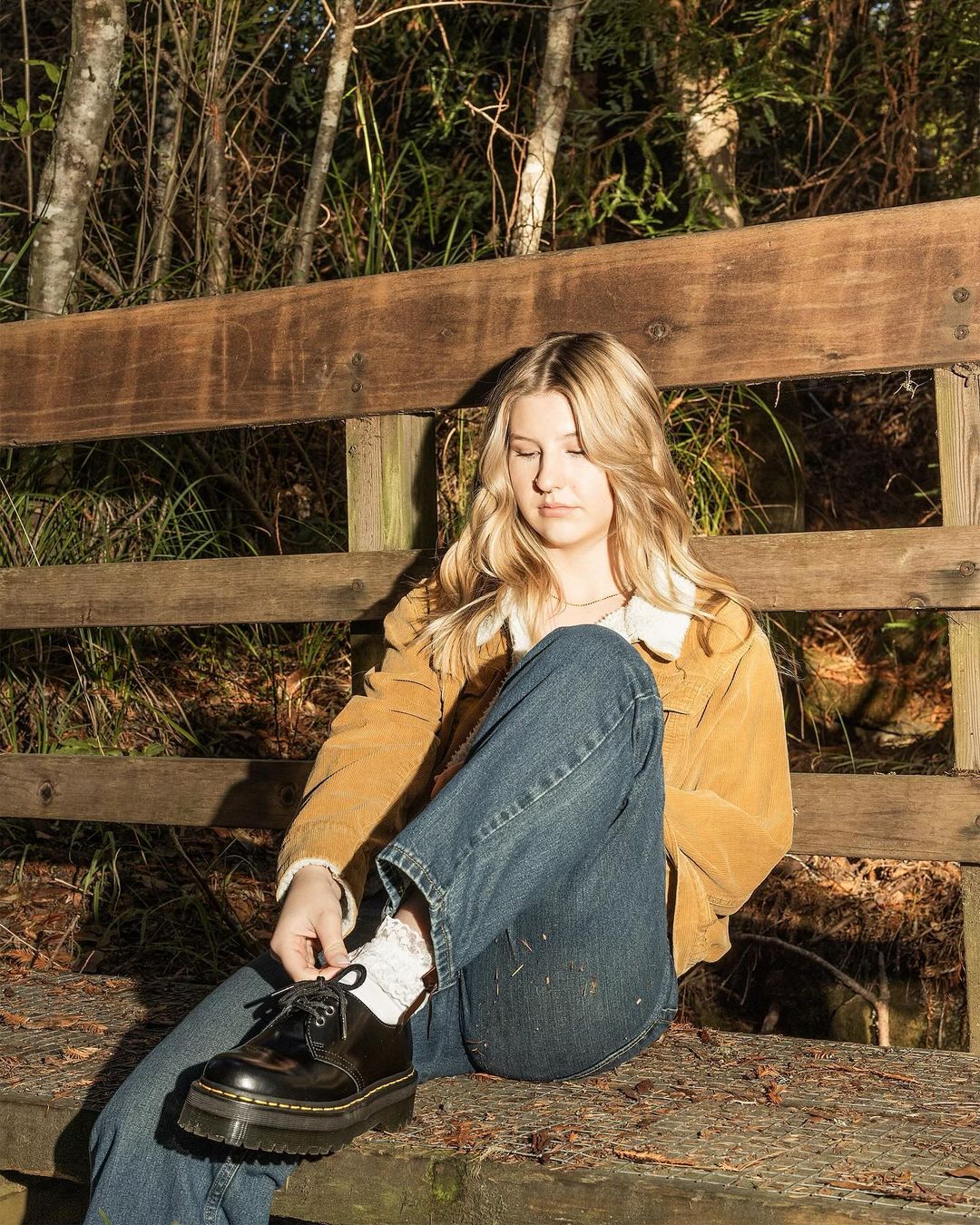 Young woman sitting on a wooden bench in a sunlit forest, tying her black platform shoe, wearing a jacket, jeans, and patterned socks.