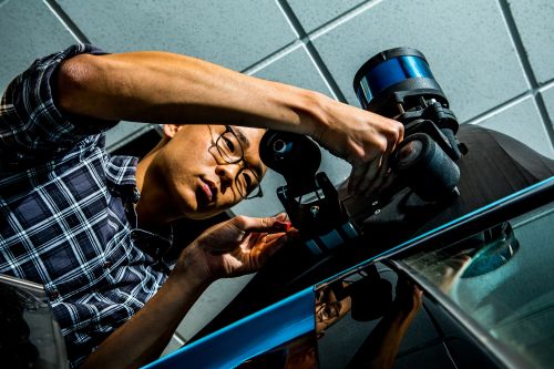 Man in plaid shirt adjusts a sensor device mounted on a dark surface, working in an indoor setting under bright lighting.