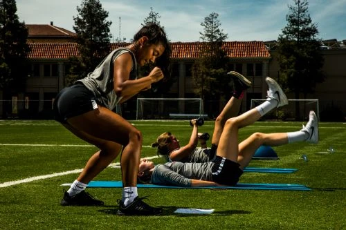 Three women in athletic wear exercise outdoors on a grassy field with building background. Woman squats as other two exercise with weights.
