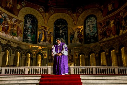Man in purple robes stands in an ornate church with murals. Interior arches and columns frame a religious figure giving a sermon.