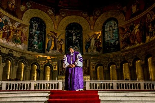 Man in purple robes stands in an ornate church with murals. Interior arches and columns frame a religious figure giving a sermon.