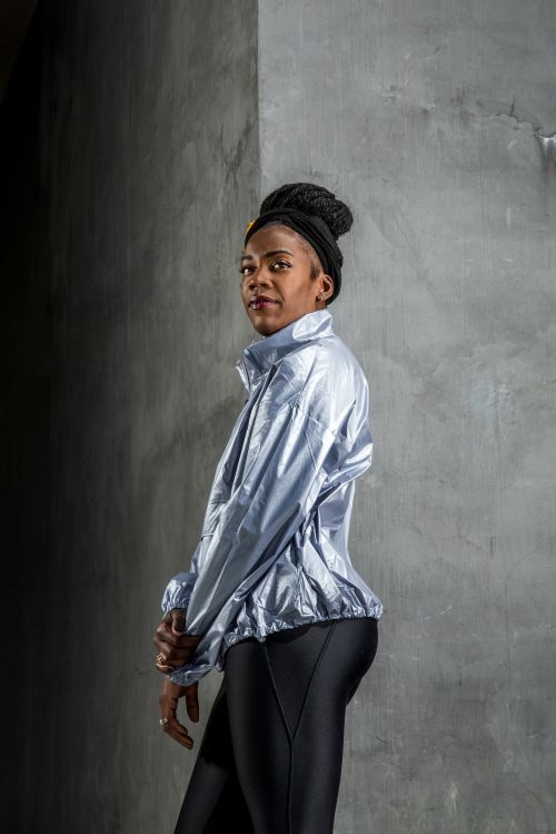 Woman in workout clothes stands against concrete wall in a medium shot, wearing a shiny light blue jacket and black leggings.