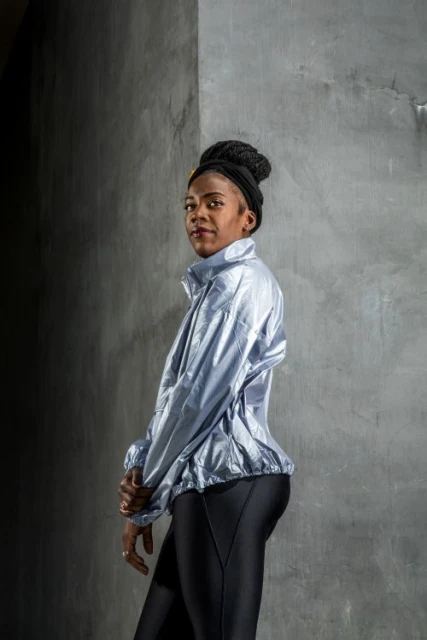 Woman in workout clothes stands against concrete wall in a medium shot, wearing a shiny light blue jacket and black leggings.