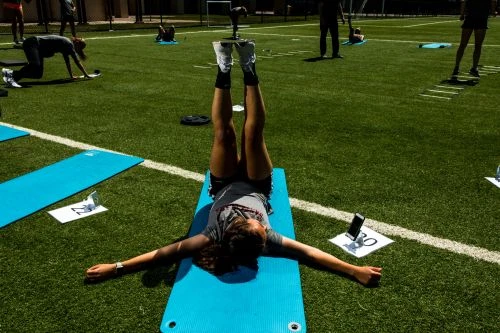 Woman lying on a blue yoga mat outdoors on green turf with legs raised in the air, with other individuals exercising in the background.