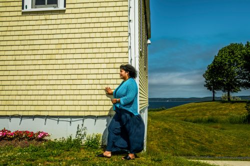 Woman walks outside yellow house wearing turquoise top and navy pants on grassy lawn with tree, ocean, and blue sky background.